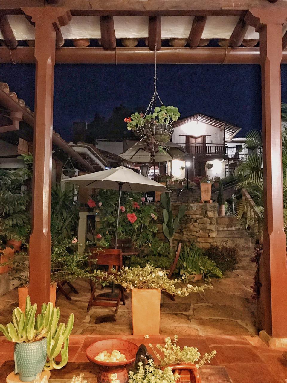 Patio interior de noche visto bajo un corredor de madera, con sombrilla, matas y flores.